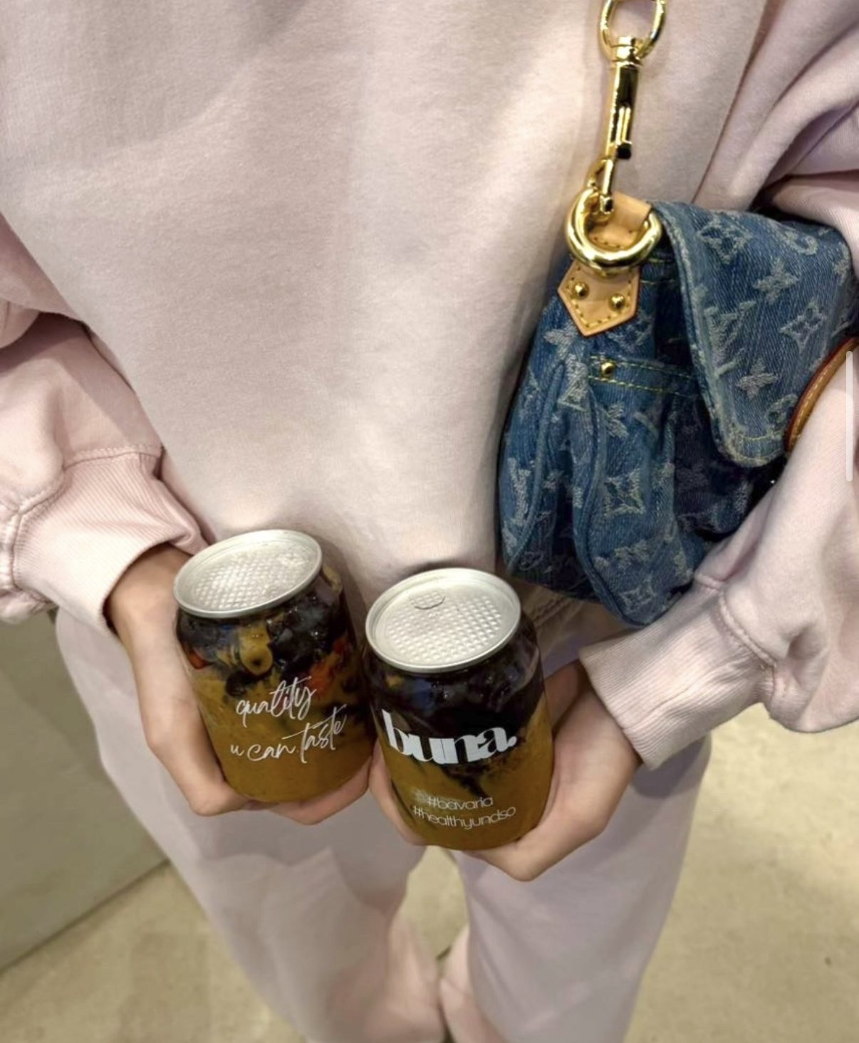 Zwei buna-Dosen mit Açaí und Erdnussbutter in den Händen, rosa Pullover, Jeanstasche